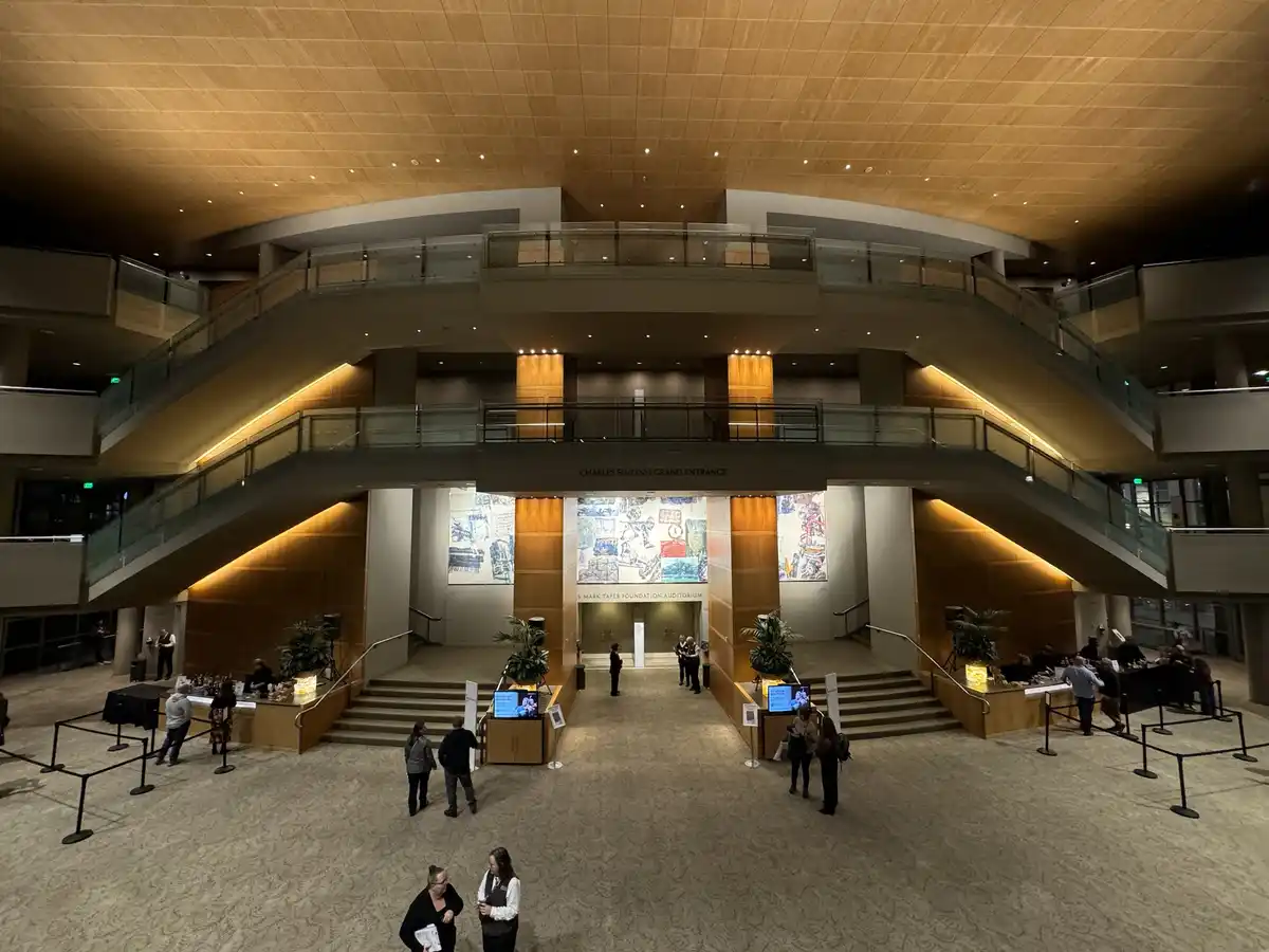 A wide-angle photo of a grand, modern building lobby with sweeping curved staircases, wooden paneling, and a high ceiling…