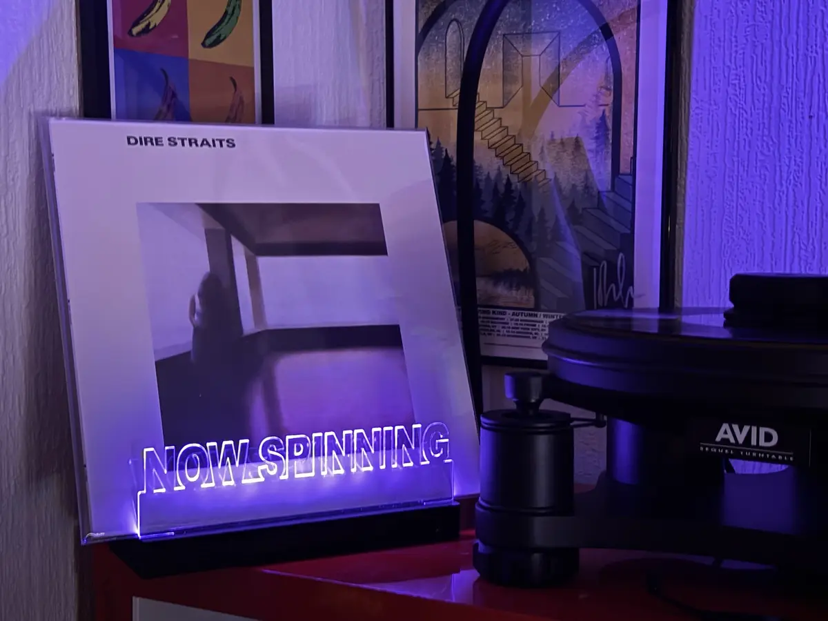 A Dire Straits album cover for 'Brothers in Arms' is propped up next to an AVID Sequel turntable, with a glowing neon sign…