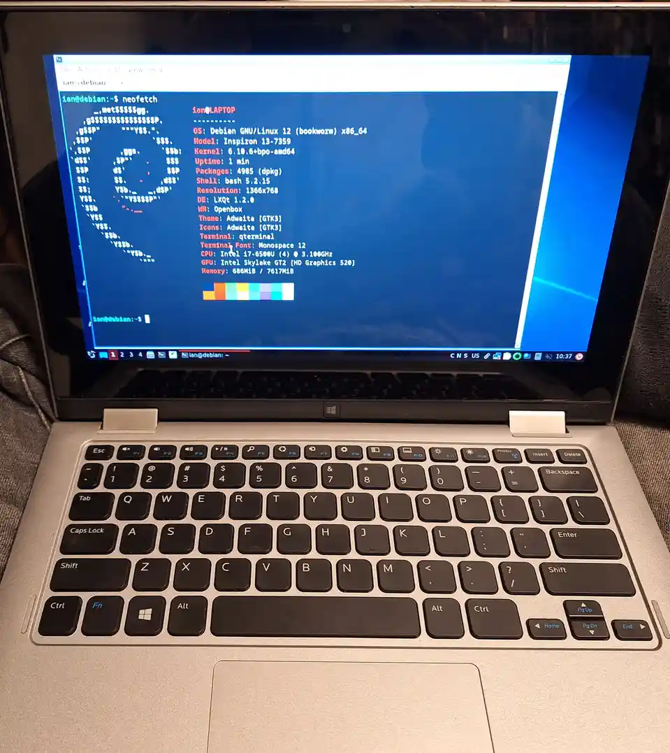 A silver laptop with a black keyboard displays a terminal window running 'neofetch' on Debian GNU/Linux 12, showing system…