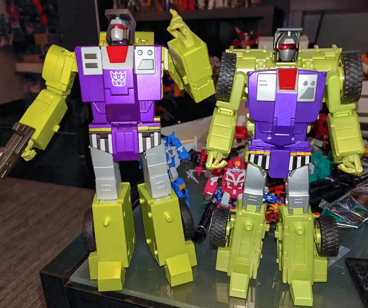 Two nearly identical large yellow and purple Transformer action figures stand side by side on a glass table, one with its…