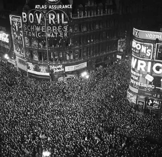 a black-and-white historical photograph of a massive, densely packed crowd filling a city street corner at night, surrounded…