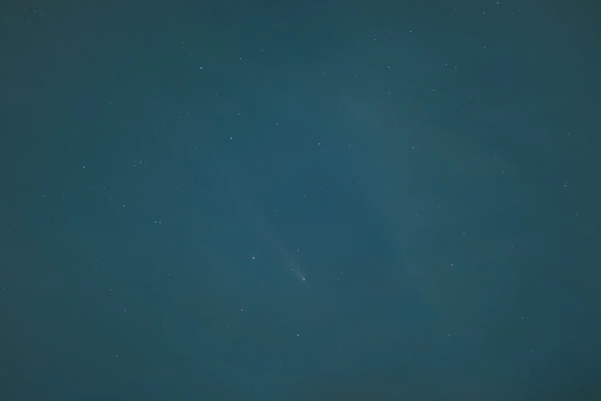 A dark teal night sky filled with faint stars, featuring a single comet with a bright nucleus and a diffuse, curved tail…