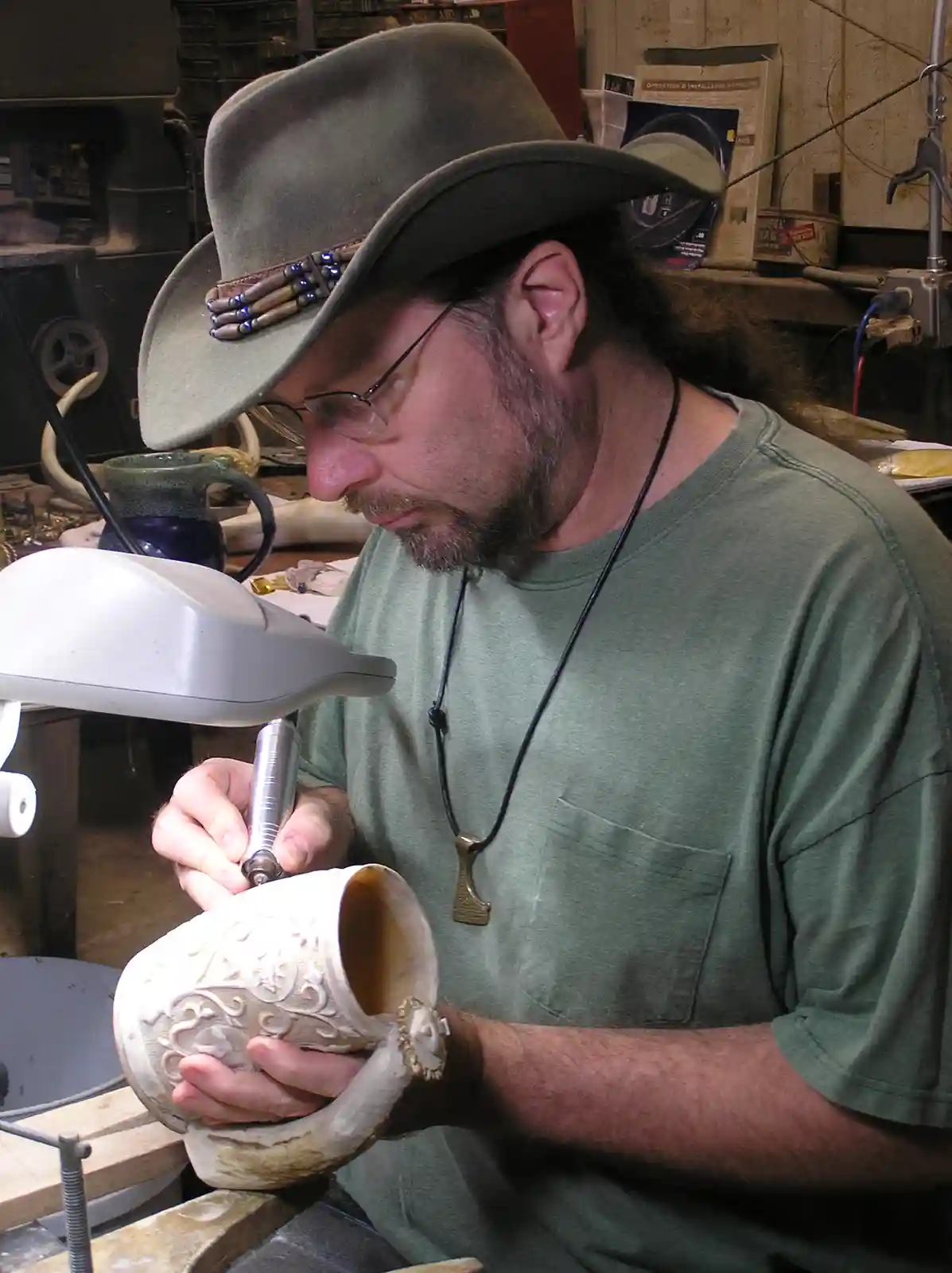 A man with long hair, glasses, and a decorated cowboy hat is intently carving intricate designs into a ceramic mug in a…
