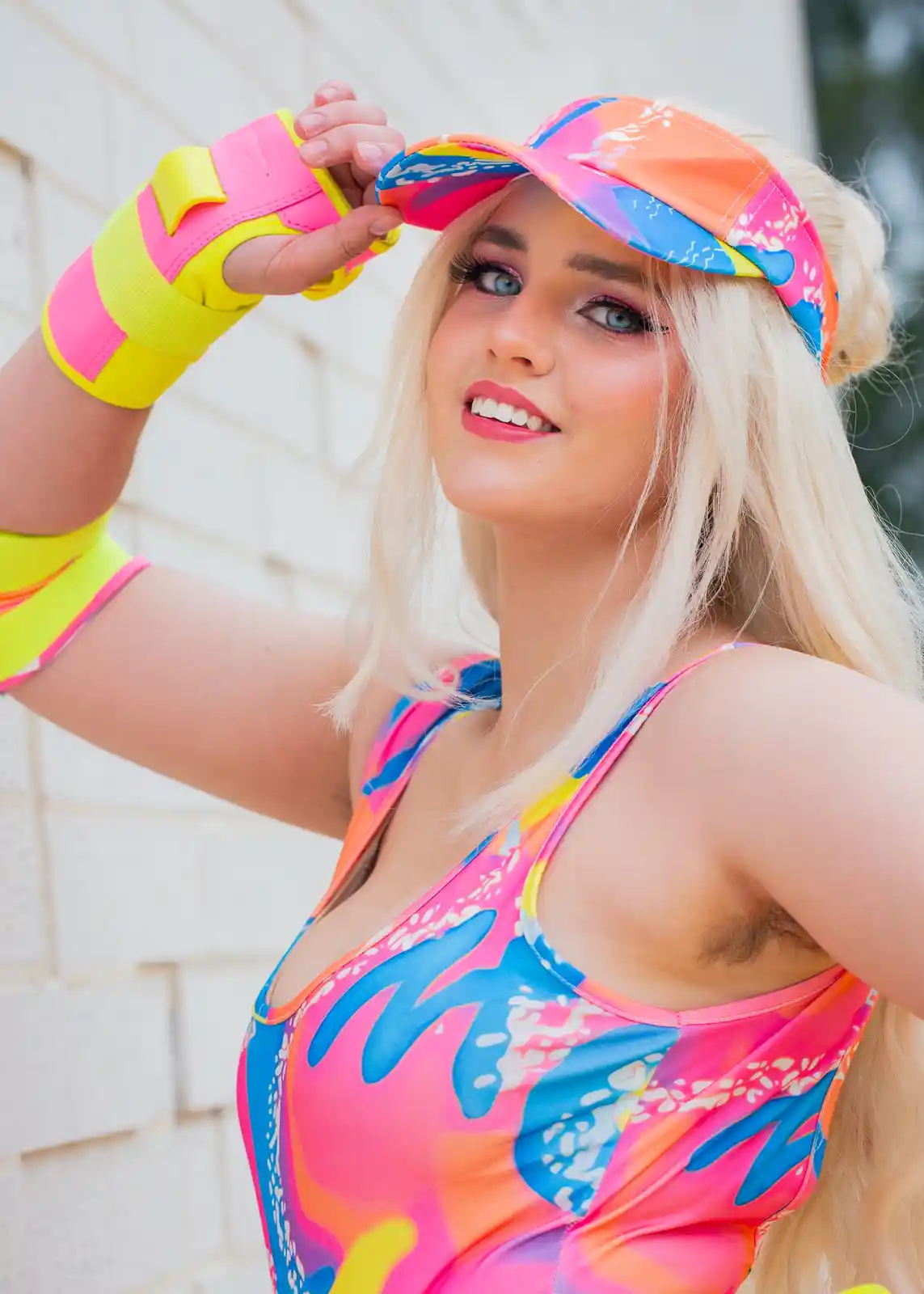 A person with blonde hair and bright makeup poses in a neon-colored, 80s-style outfit with a matching visor and arm bands…