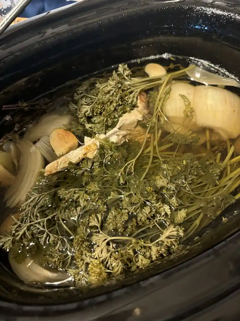 A close-up photo shows a black slow cooker filled with a simmering broth containing chicken pieces, sliced onions, garlic…