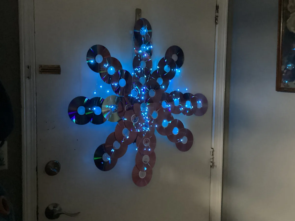 A handmade snowflake decoration made from numerous CDs and DVDs, strung together with blue fairy lights, is mounted on a…