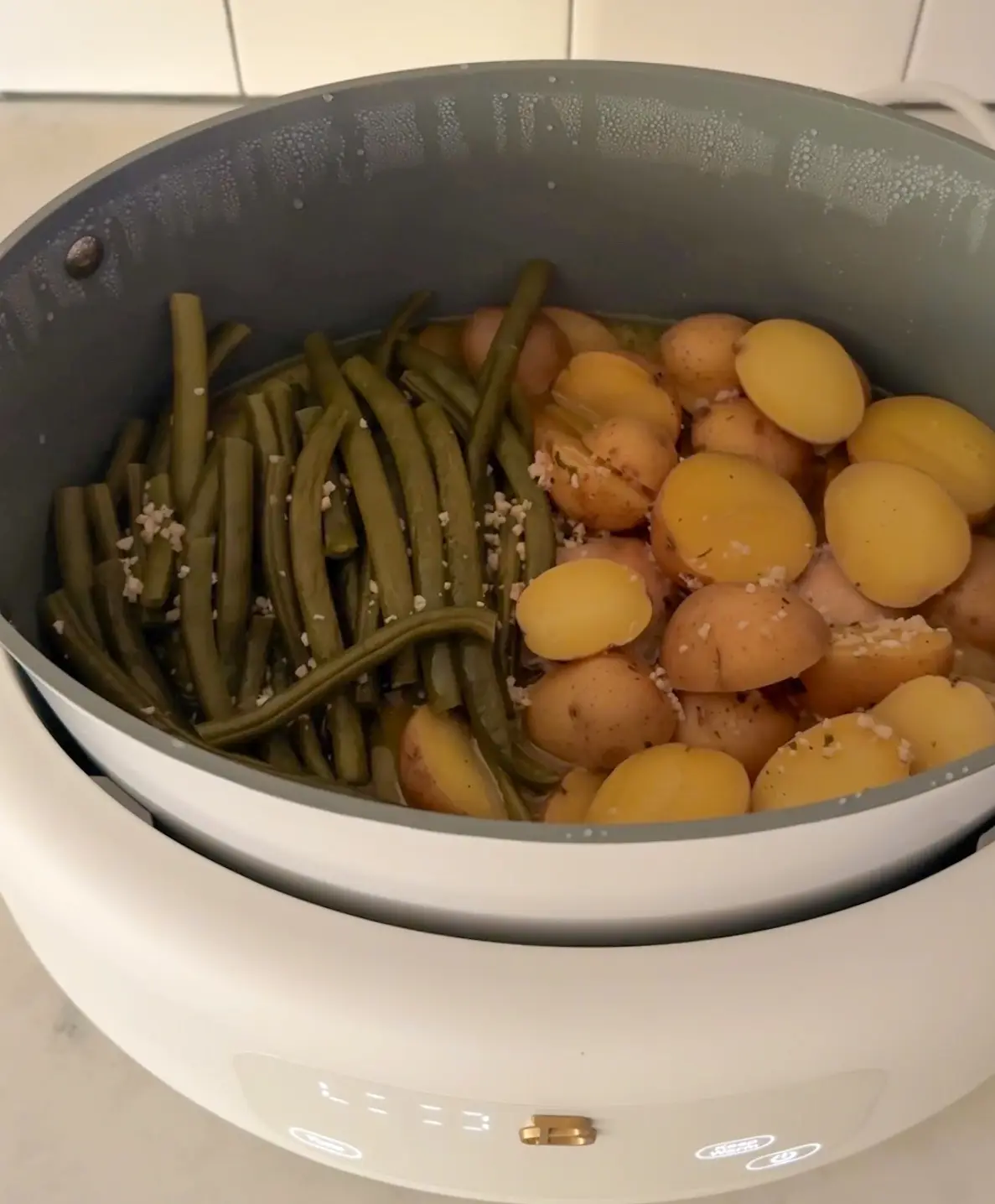 A close-up photo shows a white electric cooker with a grey inner pot containing cooked green beans and halved baby potatoes…