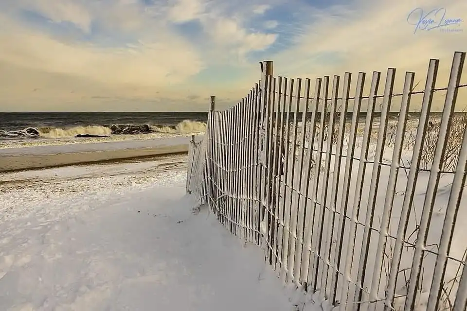 A snowy beach scene with a wooden fence running along the shore, waves crashing in the background under a cloudy sky. The…