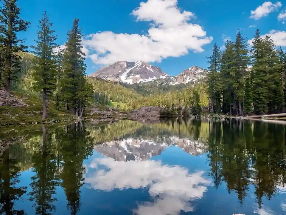 A serene landscape photo shows a calm lake perfectly reflecting snow-capped mountains and a blue sky with white clouds…