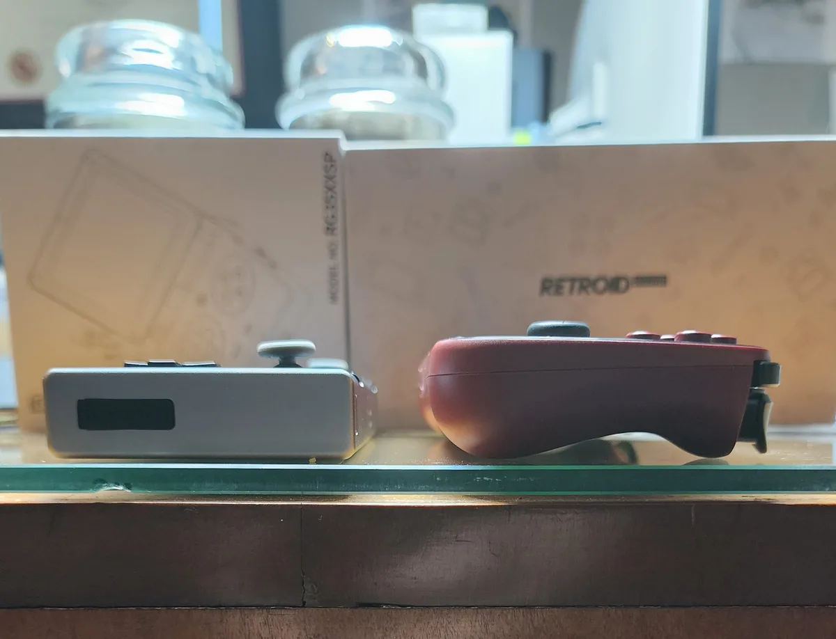 Two handheld gaming devices sit side-by-side on a glass shelf, one silver and one red, with their respective boxes visible…