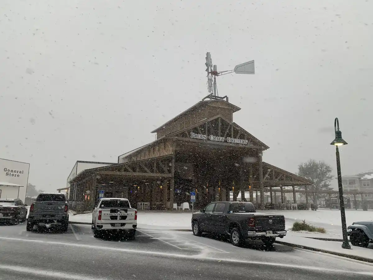 A snowy day at Props Craft Brewery, a large wooden building with a windmill on top, surrounded by parked trucks and falling…