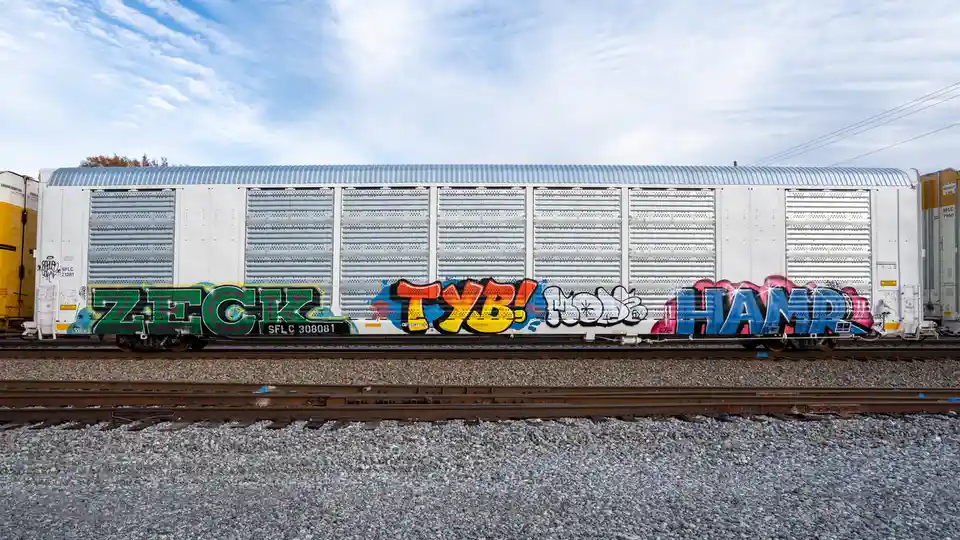 A white freight train car with corrugated metal siding sits on railway tracks under a partly cloudy sky. The side of the car…