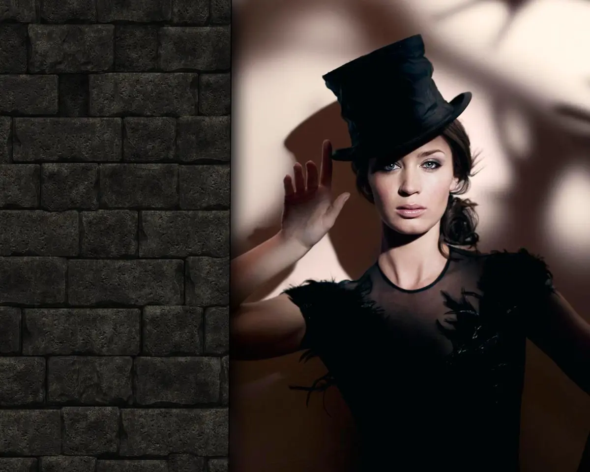 A woman in a black top hat and feathered black dress stands against a light-colored wall with dramatic shadows, her hand…