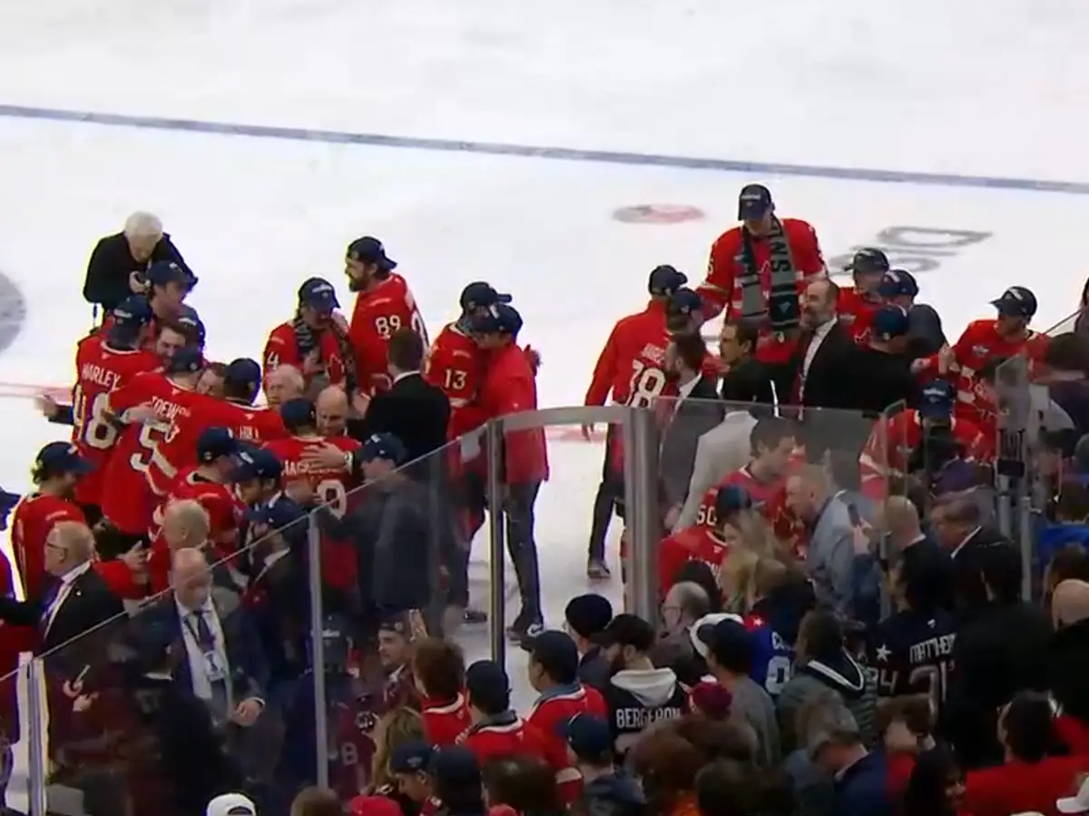 A chaotic scene at an ice hockey rink where players in red jerseys are huddled together near the boards, some jumping on…