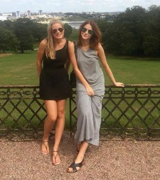 Two women stand side by side on a gravel path next to an ornate metal fence, smiling at the camera. The woman on the left…