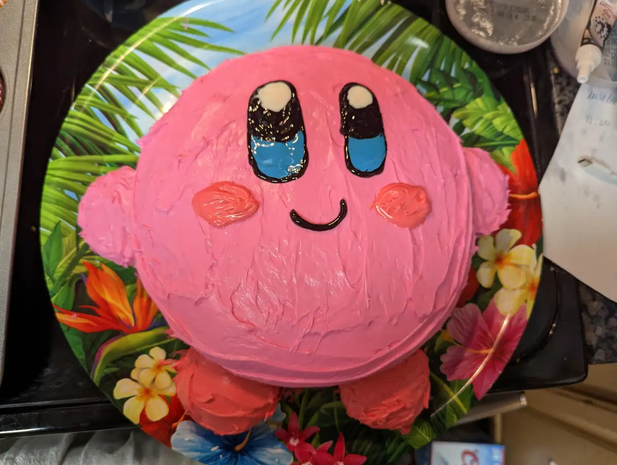 A round pink cake decorated to resemble the video game character Kirby, featuring large blue eyes with white pupils, pink…