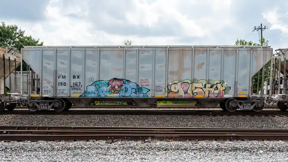 A weathered silver freight train car sits on tracks under a cloudy sky, covered in colorful graffiti including a cartoonish…