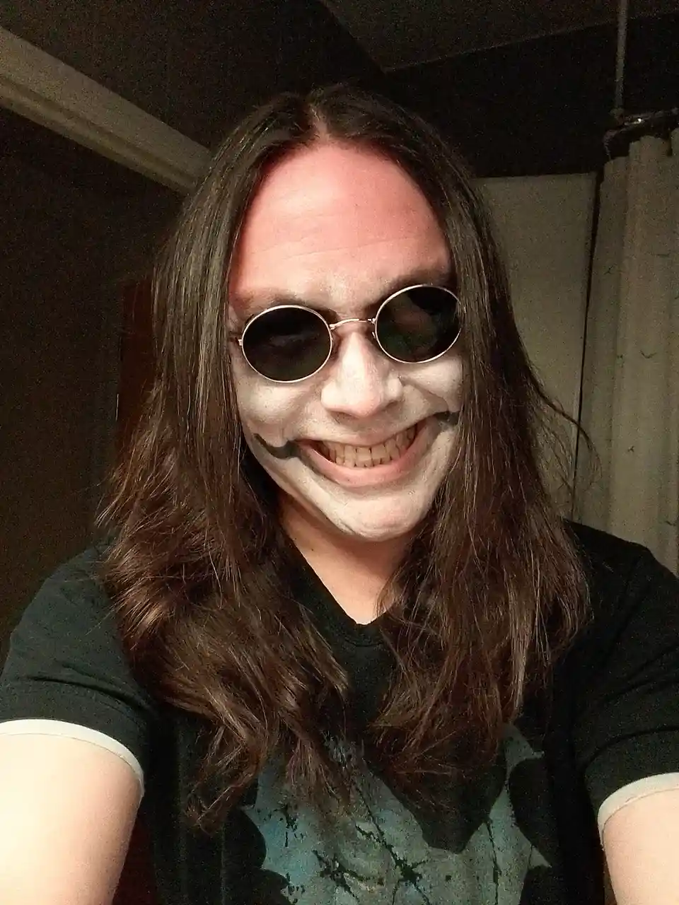 A person with long brown hair and round sunglasses has their face painted white with black makeup around their mouth to…