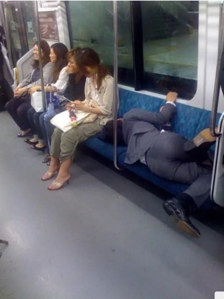A man in a suit is sleeping sprawled across a subway bench, his legs dangling off the side, while several women sit upright…