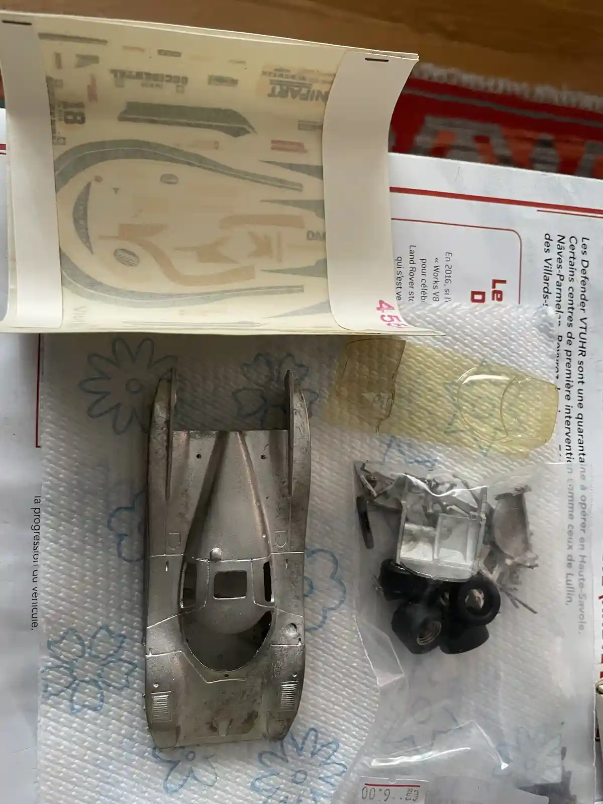 An overhead photo shows unassembled parts of a model car kit, including a large silver plastic body, small black wheels, and…