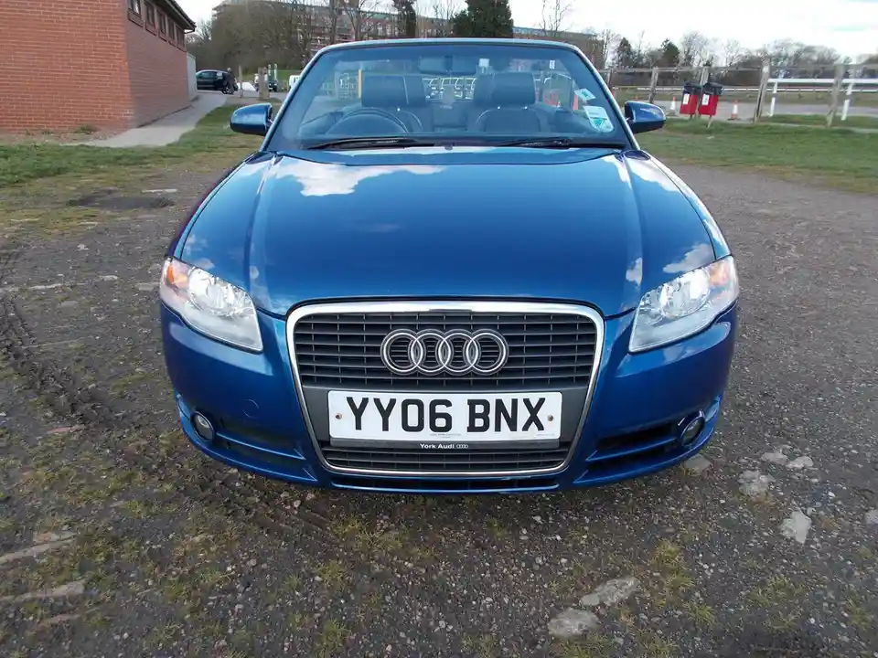 A front-facing photo of a blue Audi convertible parked on gravel, showing its grille, headlights, and license plate reading…