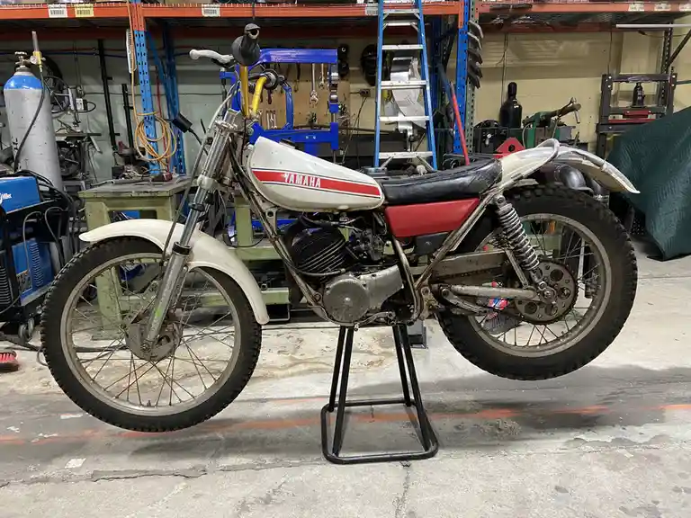 A vintage white and red Yamaha dirt bike is shown in a cluttered workshop, resting on a black stand with tools and equipment…