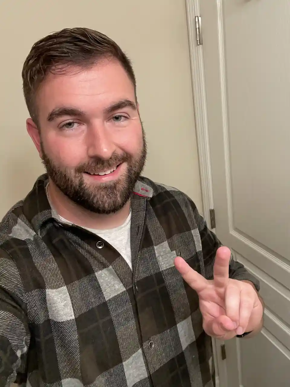 a smiling man with a beard wearing a plaid shirt over a t-shirt poses for a selfie, making a peace sign with his right hand…