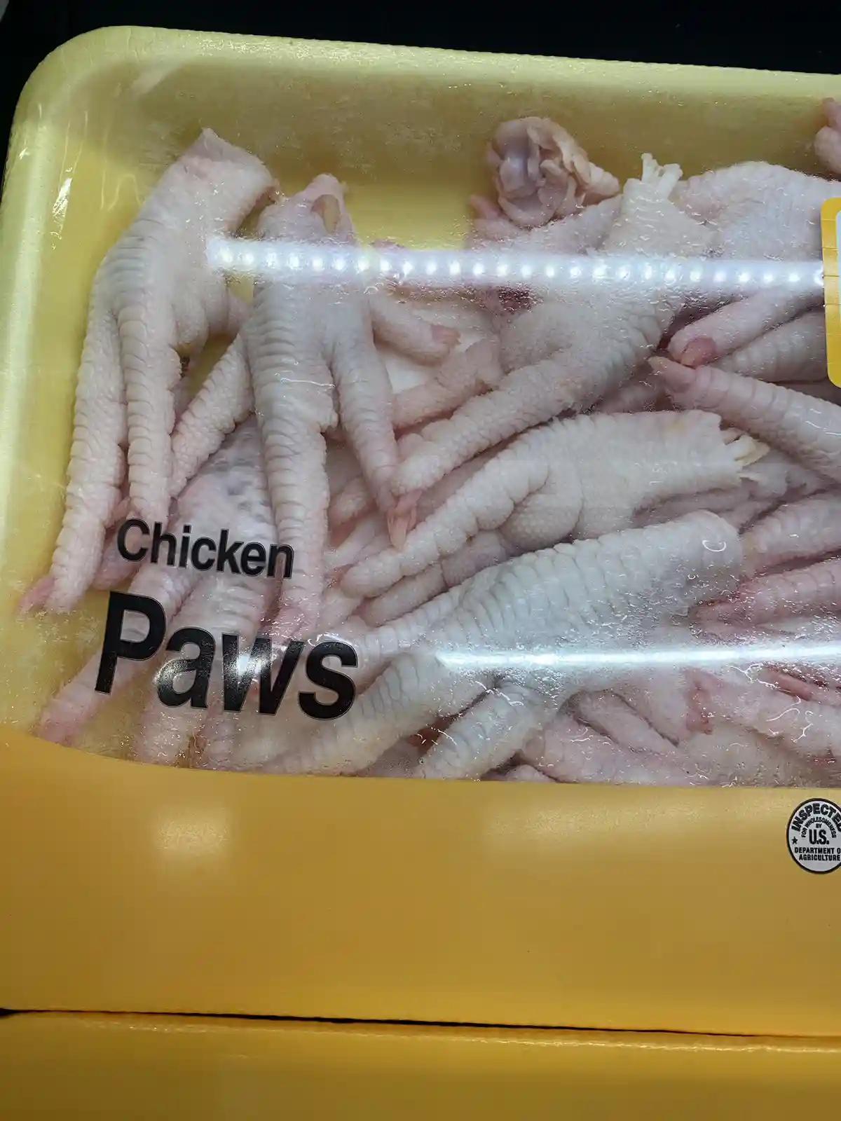 A close-up photo of a yellow plastic supermarket tray filled with raw chicken feet, sealed in plastic wrap. The label on the…