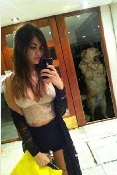 A woman in a lace top and black skirt takes a mirror selfie holding a phone and a yellow bag, while a golden retriever…