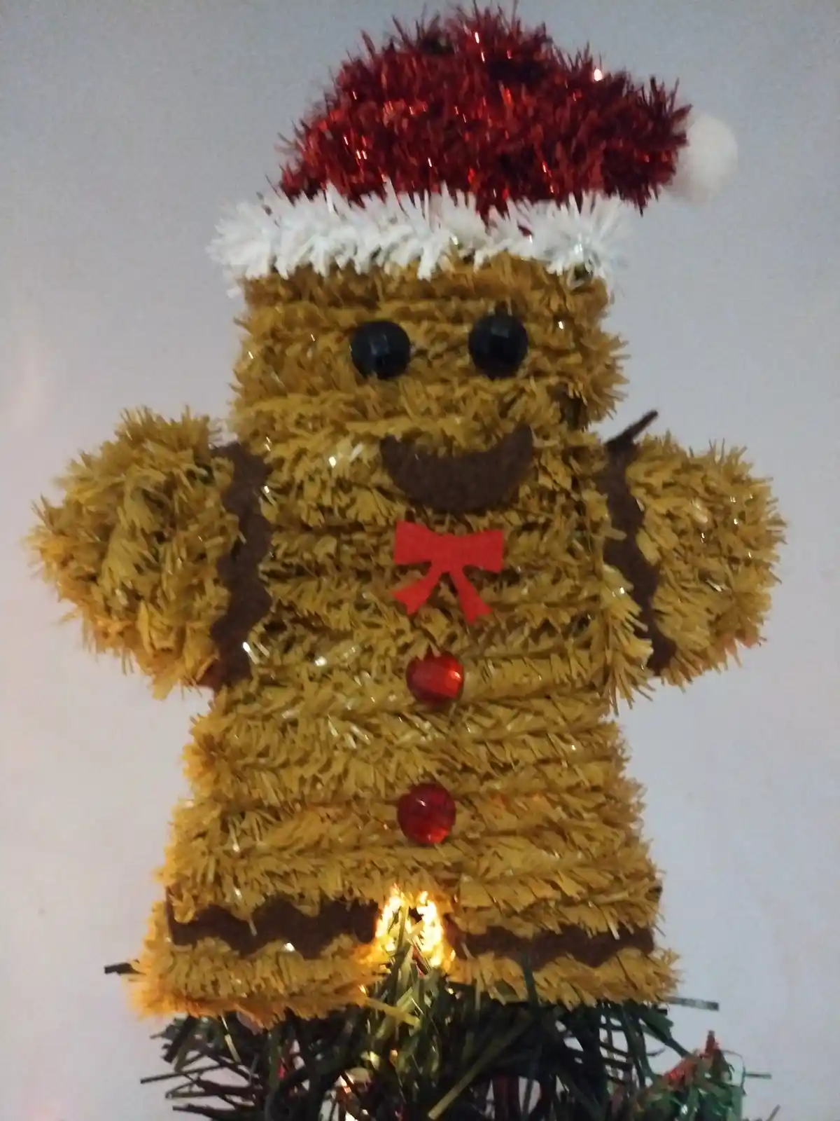 A handmade gingerbread man decoration made of gold tinsel with black button eyes, a brown smile, a red bow tie, and red…