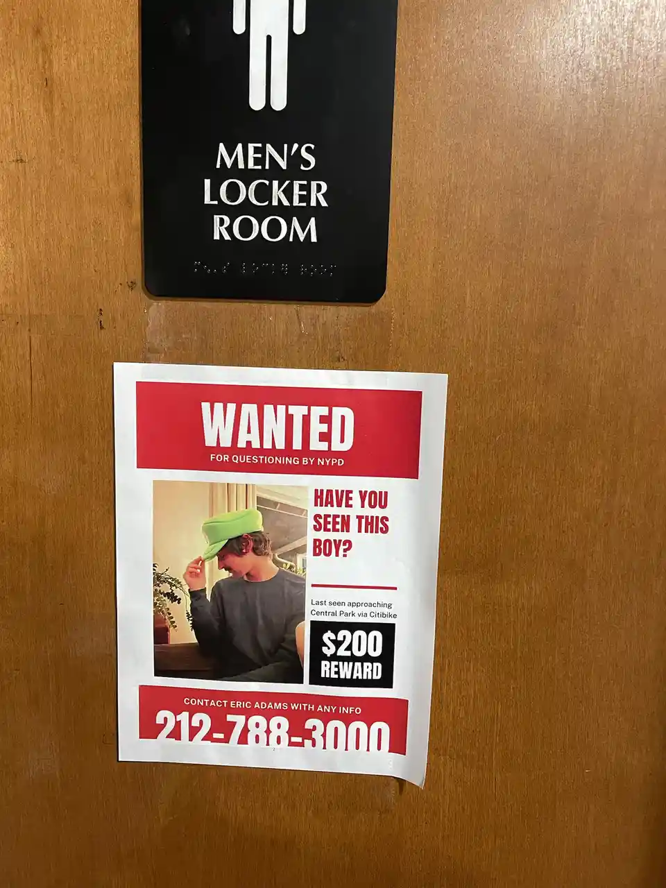 A 'WANTED' poster styled like an NYPD missing person flyer is taped to a wooden door below a 'MEN'S LOCKER ROOM' sign. The…