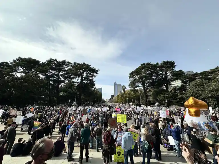 A wide-angle photo shows a large crowd of protesters gathered in a city park under a partly cloudy sky, holding numerous…