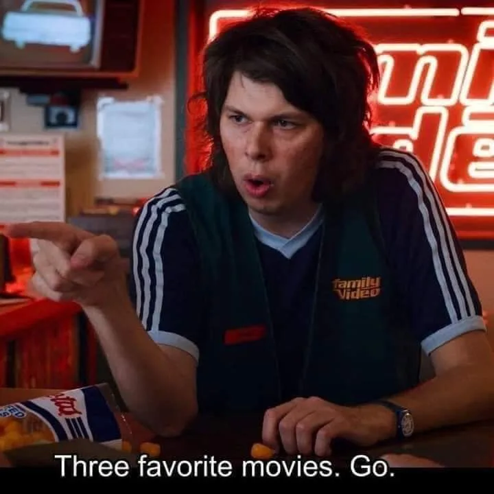 A man with shaggy brown hair wearing a green 'Family Video' vest over a dark blue shirt sits at a counter, pointing…