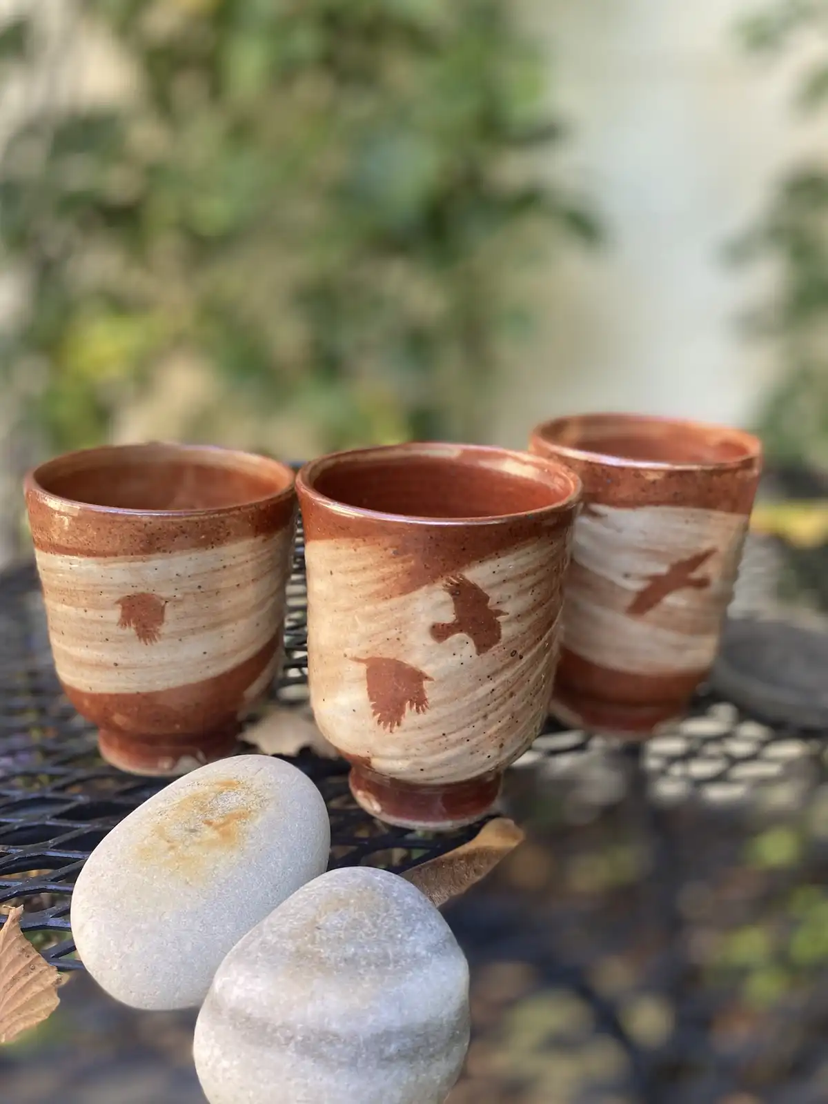Three handmade ceramic cups with brown and beige glaze and bird silhouettes sit on a black metal table outdoors, accompanied…
