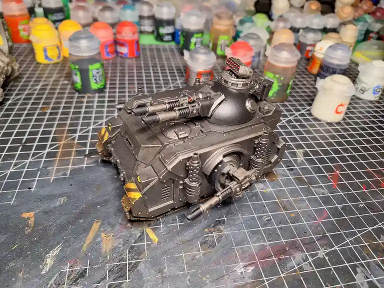 A heavily detailed, black-painted miniature tank model sits on a gridded, paint-splattered work surface, surrounded by…