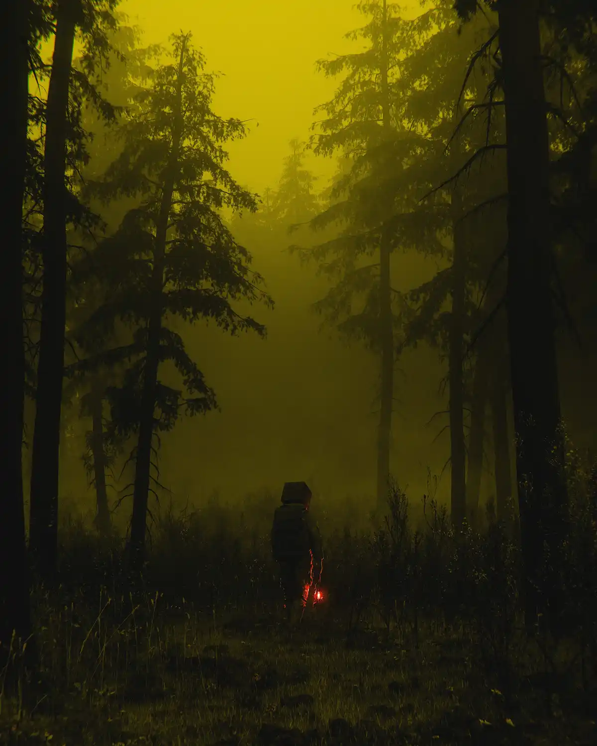 A person wearing a box-like helmet and glowing red gear walks alone through a dense, foggy forest bathed in eerie yellow…