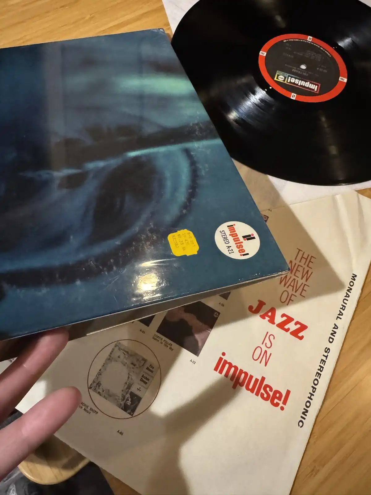 A hand holds a blue Impulse! Records vinyl album sleeve next to a black vinyl record and a promotional flyer. The flyer…