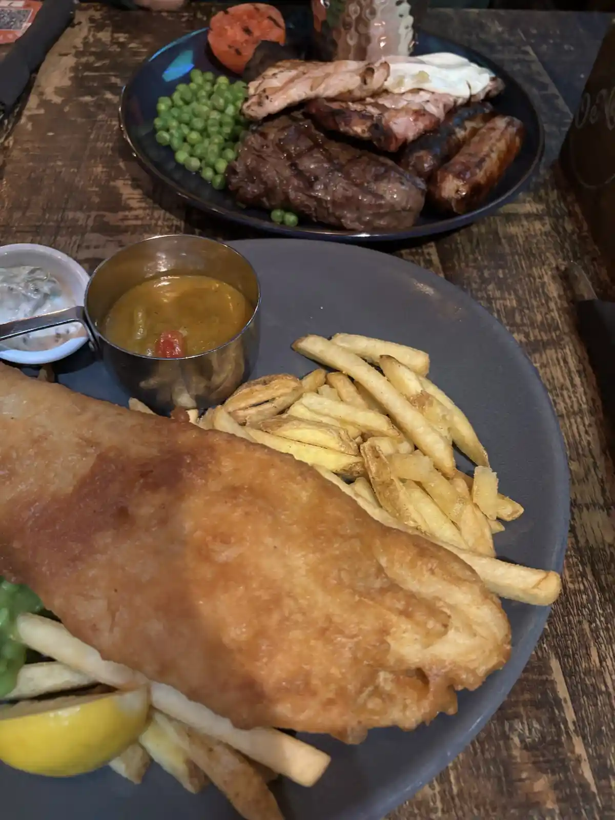 A close-up photo shows a plate of fish and chips in the foreground, with a full english breakfast (steak, sausages, bacon…