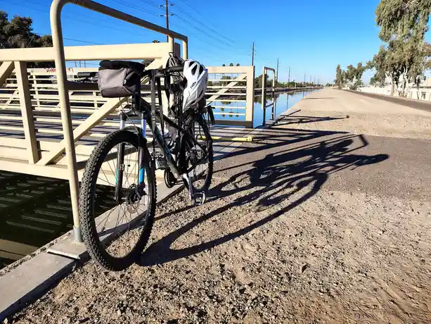 A black mountain bike with a white helmet and a grey bag is parked on a gravel path next to a canal, leaning against a beige…