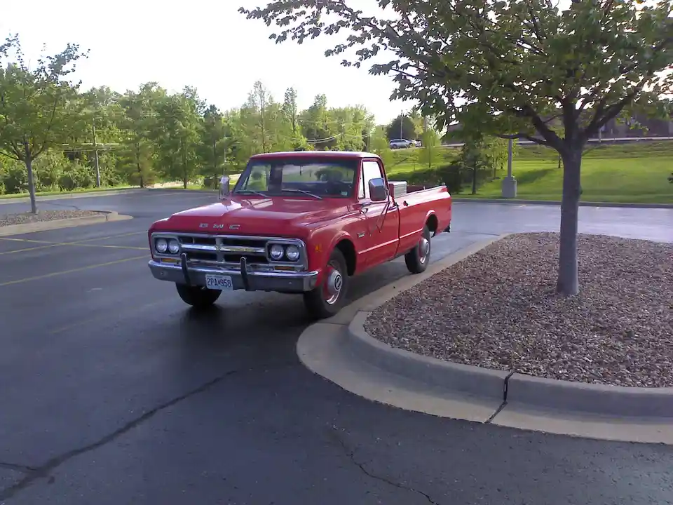 A red vintage GMC pickup truck is parked at an angle in a paved parking lot next to a tree in a gravel bed. The truck has…