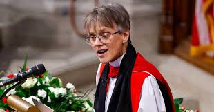 A woman in academic robes and glasses stands at a podium with flowers, looking slightly nervous while speaking into a…