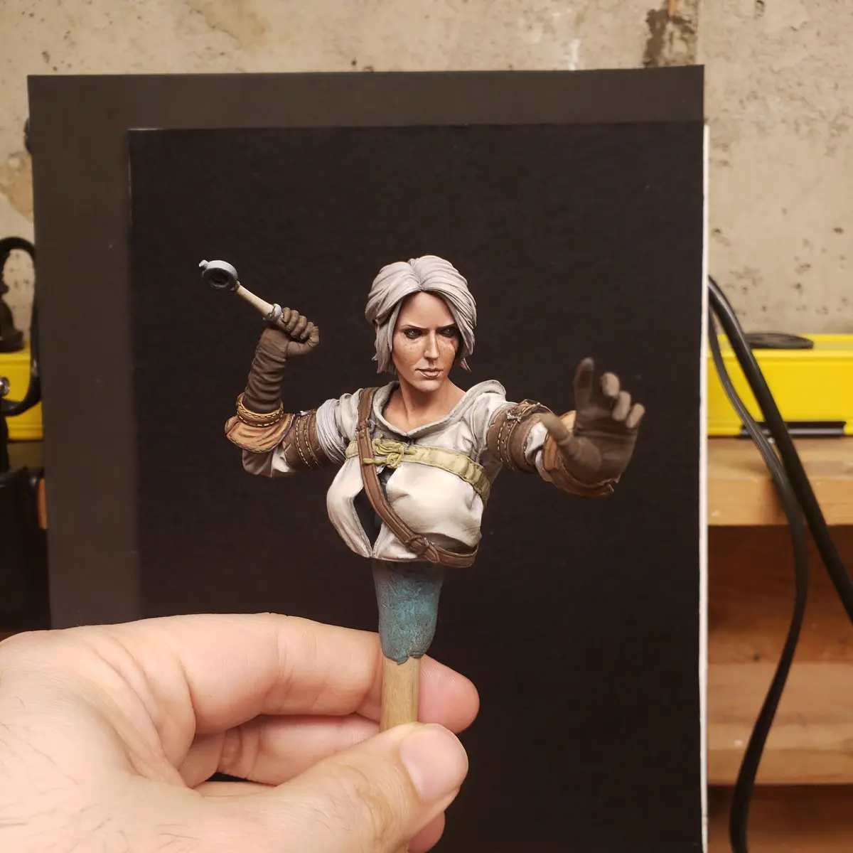 A hand holds a highly detailed miniature bust of a female fantasy character with grey hair, holding a small hammer in one…