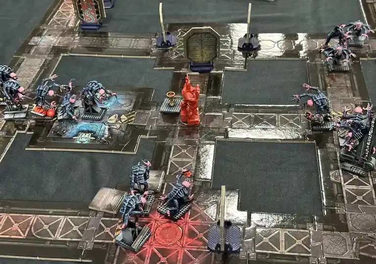 A tabletop wargame scene shows numerous blue and pink miniatures surrounding a single red miniature, which is centrally…