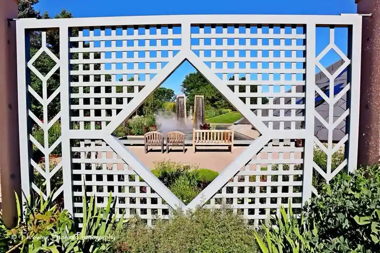 A white lattice garden fence with a diamond-shaped opening frames a view of a landscaped garden with benches and water…