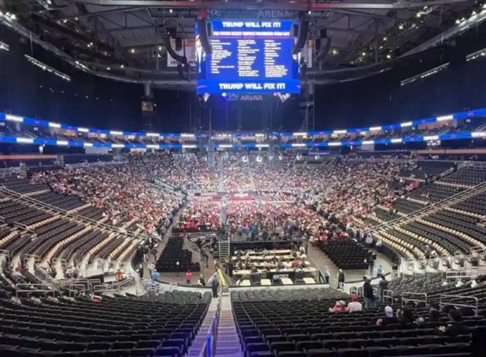 A wide-angle photo of a large, mostly empty arena with a few scattered people in the stands. The central focus is a massive…