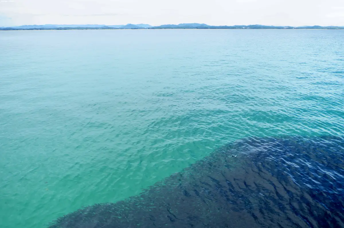 A wide shot of calm, turquoise ocean water meeting a darker, deeper blue section near the foreground, with distant mountains…