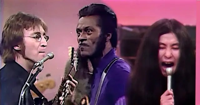 A still from a 1960s TV show featuring three musicians: John Lennon on the left holding a microphone, a Black man in a…