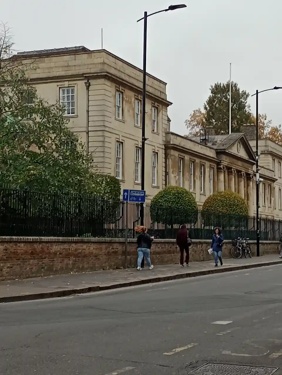 A street scene with pedestrians walking past a large stone building behind a fence, with a blue bus and bike sign visible…