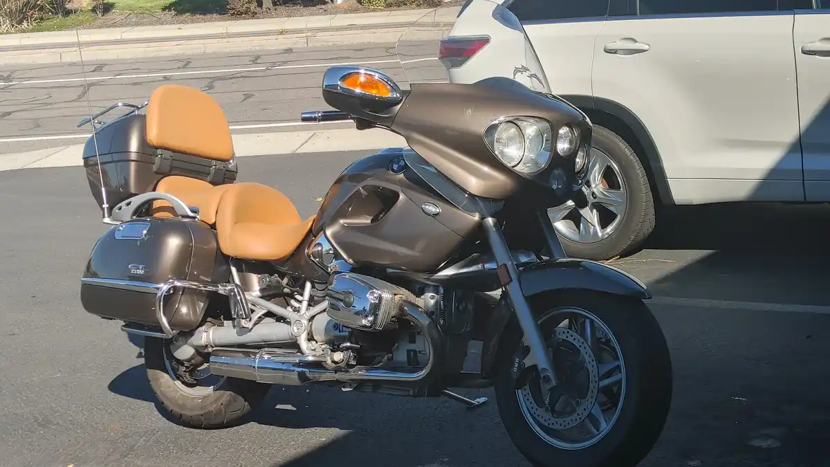 A brown BMW motorcycle with tan leather seats and chrome accents is parked next to a white SUV on an asphalt road. The…