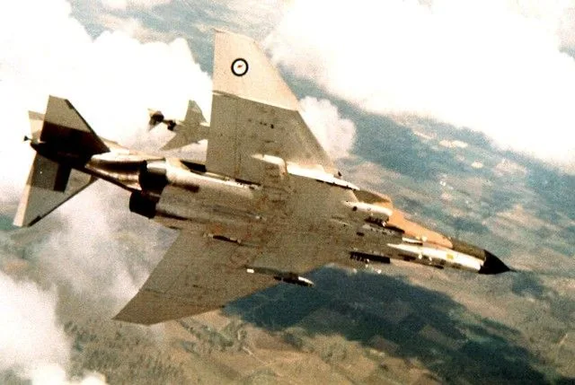 A military fighter jet, likely a British RAF aircraft with a roundel insignia on its tail, is shown flying above clouds and…